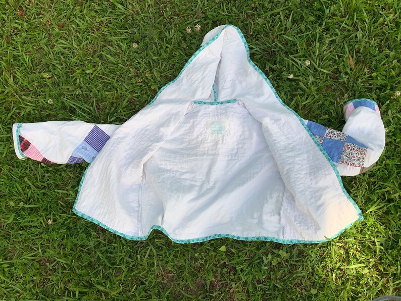 May include: A white quilted jacket with a hood. The jacket is made of patchwork fabric with different colored squares. The jacket is lined with a white fabric and has a turquoise trim.