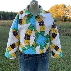 May include: A white quilted jacket with a patchwork design featuring brown, yellow, green, and blue fabric. The jacket has a button closure and long sleeves.