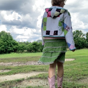 May include: A person wearing a white quilted jacket with a green and pink patchwork design, a green tiered skirt, and pink cowboy boots. The person is standing in a grassy field with a cloudy sky in the background.
