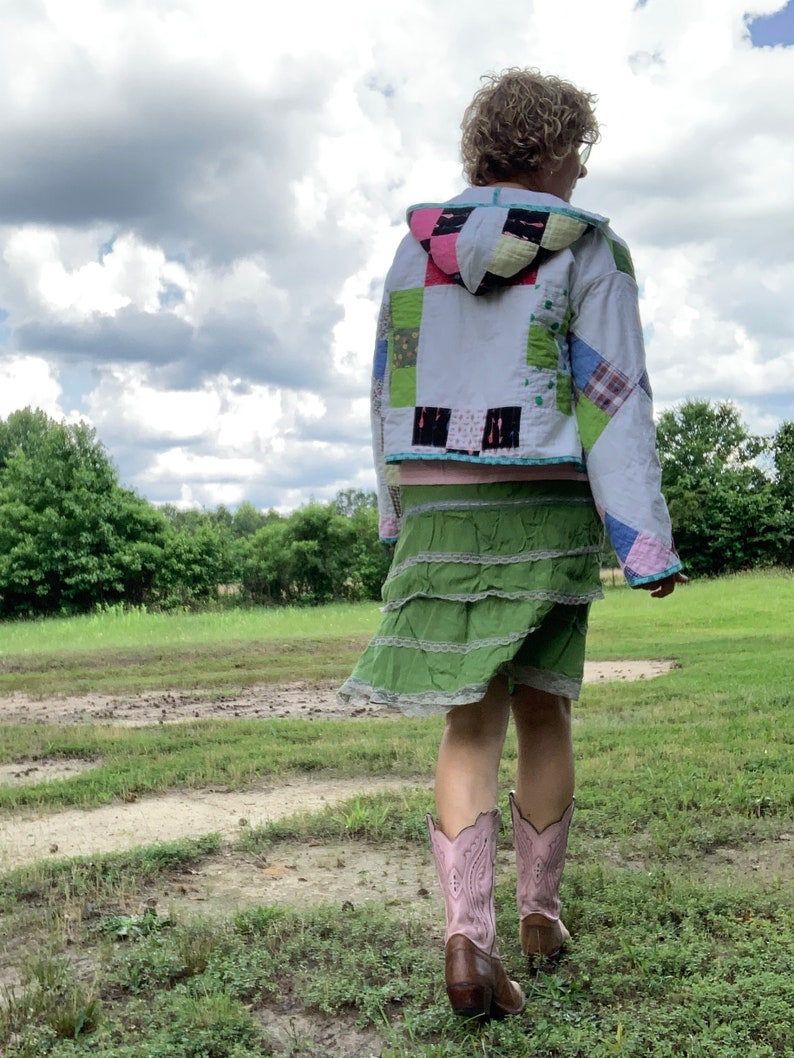May include: A person wearing a white quilted jacket with a green and pink patchwork design, a green tiered skirt, and pink cowboy boots. The person is standing in a grassy field with a cloudy sky in the background.