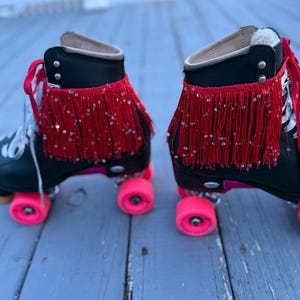 May include: Pair of black roller skates with bright pink wheels. The skates have red fringe with silver sequins around the ankles. The laces are white and red. The skates are on a weathered gray wooden surface.
