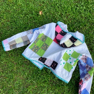 May include: A white quilted jacket with a patchwork design. The jacket has a hood and long sleeves. The patchwork is made of different colored fabrics, including green, pink, blue, and red.