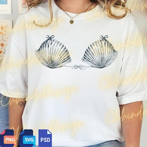 May include: White t-shirt featuring a seashell bra design. The graphic shows two seashell shapes with bow details, connected by a string. The seashells have a detailed, textured appearance with a gradient of colors. The design is centered on the shirt.