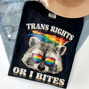 May include: Black t-shirt with the text "TRANS RIGHTS" and "OR I BITES" in white and rainbow colors. The shirt features a raccoon wearing rainbow sunglasses and holding a rainbow flag. The shirt is on top of a pair of blue jeans.