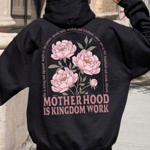 May include: Black hoodie featuring a floral design with pink peonies and green leaves. The text "MOTHERHOOD IS KINGDOM WORK" is printed below the flowers. Additional text arcs around the floral design.