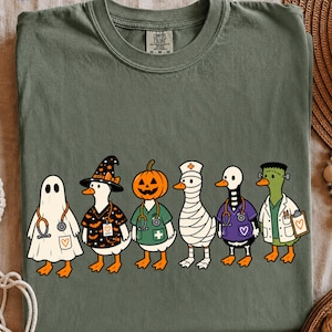 Pode incluir: Uma t-shirt verde-oliva com um gráfico de seis patos vestidos com vários trajes de Halloween, incluindo um fantasma, uma bruxa, uma abóbora, uma múmia, um esqueleto e um monstro, todos usando uniformes médicos e estetoscópios com corações.