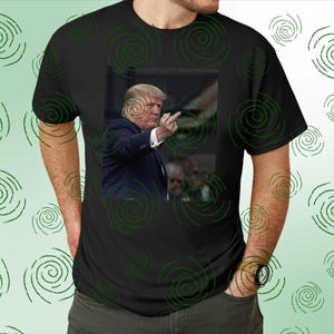 May include: Black t-shirt featuring a photographic image of a man in a suit, raising his middle finger. The shirt has a green spiral pattern. The man in the image has blonde hair and is wearing a dark suit.