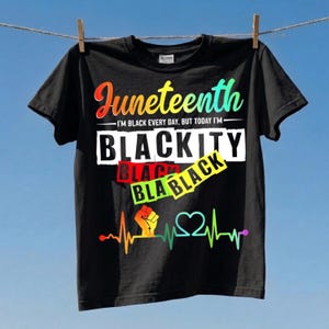 Puede incluir: Camiseta negra con el texto "Juneteenth" en colores del arcoíris. La camiseta también presenta el texto "I'm black every day, but today I'm" y la palabra "BLACKITY" en blanco. Un gráfico de latido del corazón colorido en la parte inferior.
