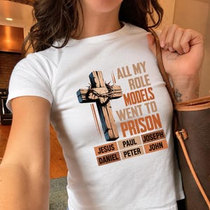 May include: A white short-sleeved t-shirt featuring a graphic of a wooden cross with a crown of thorns. The text reads ALL MY ROLE MODELS WENT TO PRISON, followed by a list of names: JESUS, PAUL, JOSEPH, DANIEL, PETER, and JOHN in orange boxes.