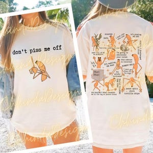 May include: Off-white t-shirt with the phrase "don't piss me off" and a cartoon illustration. The back of the shirt has multiple cartoon graphics and text, including "RIGATONI PASTA" and "sweet spicy chili tortilla chips."