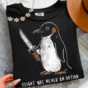 May include: Black crew neck t-shirt featuring a cartoon penguin illustration holding a knife. The penguin is black and white with a brown beak and feet. The text "FLIGHT WAS NEVER AN OPTION" is printed below the penguin.