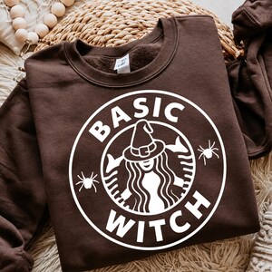 May include: A dark brown crewneck sweatshirt featuring a white circular graphic print. The design includes the text BASIC WITCH, a central figure wearing a pointed witch hat, and two small spider illustrations on the left and right sides.