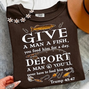 May include: A dark brown t-shirt featuring a graphic of a fish, a fishing rod, and fishing lures. The text reads: GIVE A MAN A FISH, you feed him for a day. DEPORT A MAN & YOU'LL never have to feed him again. Trump 45:47 is printed at the bottom.