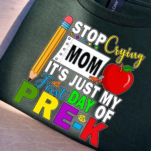 May include: A dark green t-shirt featuring a colorful graphic with a yellow pencil, a red apple, and a notebook page. The text reads: STOP CRYING MOM IT'S JUST MY FIRST DAY OF PRE-K. The design includes a small orange ruler and a yellow paint palette.