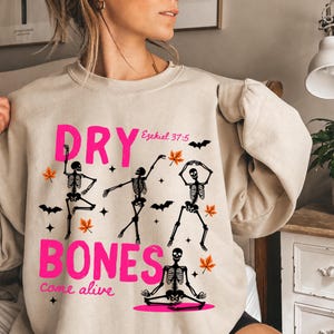 Könnte beinhalten: Ein beigefarbenes Sweatshirt mit Rundhalsausschnitt und einem Grafikdruck mit dem leuchtend pinken Text "DRY BONES" und "come alive". Das Design zeigt vier schwarze Skelettfiguren in verschiedenen Posen, orange Herbstblätter, Fledermäuse und Sterne.