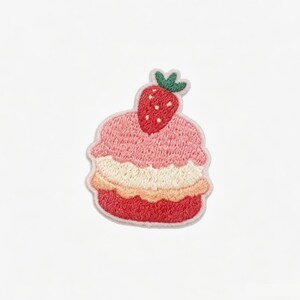 Op de afbeelding: Een geborduurde patch in de vorm van een gelaagd dessert met een rode aardbei erbovenop. Het ontwerp heeft een roze bovenlaag, een crèmekleurige tussenlaag en een rode onderlaag, allemaal omlijnd met een lichtroze rand op een witte achtergrond.