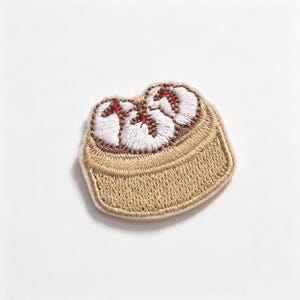 May include: An embroidered patch featuring a steamed dumpling in a bamboo steamer basket. The dumpling has a white dough exterior with red accents at the top, while the basket is rendered in shades of tan and gold thread with a textured finish.