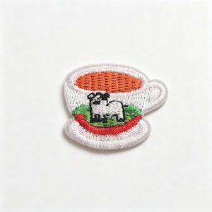 May include: An embroidered patch shaped like a white teacup with a handle. The cup contains a bright orange liquid, a patch of green grass, and a small black and white dog standing in the centre. The base of the cup is outlined in red thread.
