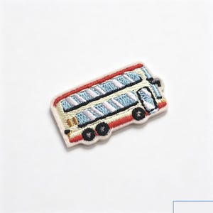 May include: An embroidered patch shaped like a double-decker bus. The bus features a cream-colored body with red trim along the top and bottom, light blue windows with white diagonal accents, and black wheels. The patch has a white fabric border.