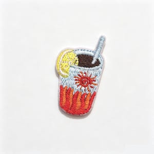 Puede incluir: Un parche bordado que representa un vaso de bebida helada con líquido marrón oscuro, una pajita azul claro y una rodaja de limón amarilla. El vaso presenta un gráfico de sol rojo y naranja y un patrón de llamas rojas en la base.