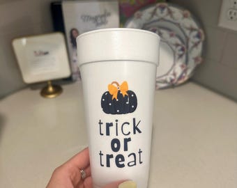 Puede incluir: Vaso de plástico blanco con un gráfico de calabaza negra y las palabras "trick or treat" en negro. La calabaza tiene un lazo naranja. El vaso está sostenido por una persona.