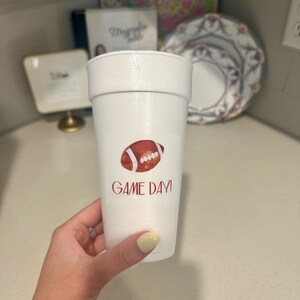 May include: A white foam cup featuring a red American football graphic and the text "GAME DAY!". The cup is held by a person. Ideal for serving drinks during a sports event.