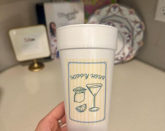 Op de afbeelding: Een witte plastic beker met een "Happy Hour" label met een cocktailglas, citroen en munt illustratie. De beker wordt vastgehouden door een hand.