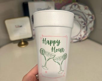 Op de afbeelding: Een witte schuimrubberen beker met de woorden "Happy Hour" in groene letters, samen met twee margarita glazen. De beker wordt door een persoon vastgehouden. De achtergrond bevat een roze en groen bloemenpatroon en een decoratief bord.