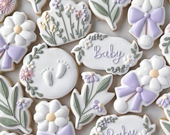 May include: A collection of decorated sugar cookies featuring white icing with pastel purple and sage green accents. Designs include daisies with purple bows, floral sprigs, baby footprints, and plaques with the word Baby written in script.
