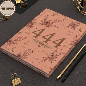 May include: A hardcover journal with a dusty rose cover featuring a repeating pattern of the number 444, floral illustrations, and crescent moons. The central design highlights the number 444 in a large, bold font with a moth graphic underneath.