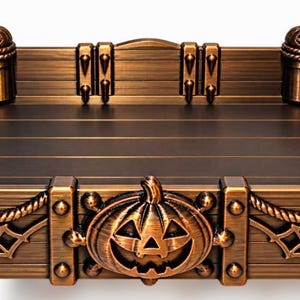 May include: A rectangular tray with a metallic bronze finish, featuring four corner posts topped with carved jack-o'-lanterns. The sides are decorated with spider web motifs, rope-like trim, and a central jack-o'-lantern on the front panel.