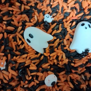 May include: A mixture of orange and black dyed rice grains scattered with several small white ghost-shaped figurines, a tiny white ghost, and a single white round eyeball-shaped piece with a black pupil and a small red spot.