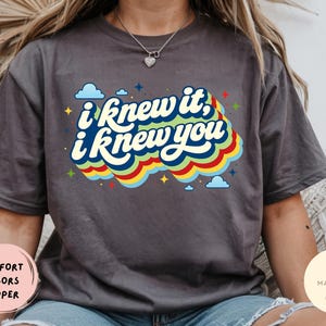 May include: A charcoal gray short-sleeve t-shirt featuring the text "i knew it, i knew you" in a bold, layered font. The graphic includes a colorful, wavy rainbow-inspired background with small blue clouds and yellow star accents surrounding the text.