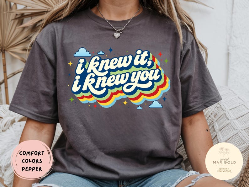 May include: A charcoal gray short-sleeve t-shirt featuring the text "i knew it, i knew you" in a bold, layered font. The graphic includes a colorful, wavy rainbow-inspired background with small blue clouds and yellow star accents surrounding the text.