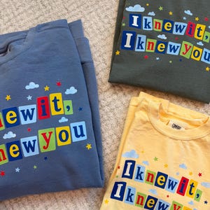 Op de afbeelding: Drie shirts met ronde hals in blauw, olijfgroen en geel. Elk shirt heeft een grafische print met de tekst "I knew it, I knew you" in kleurrijke blokletters, omringd door kleine blauwe wolken en rode, gele en blauwe sterren.
