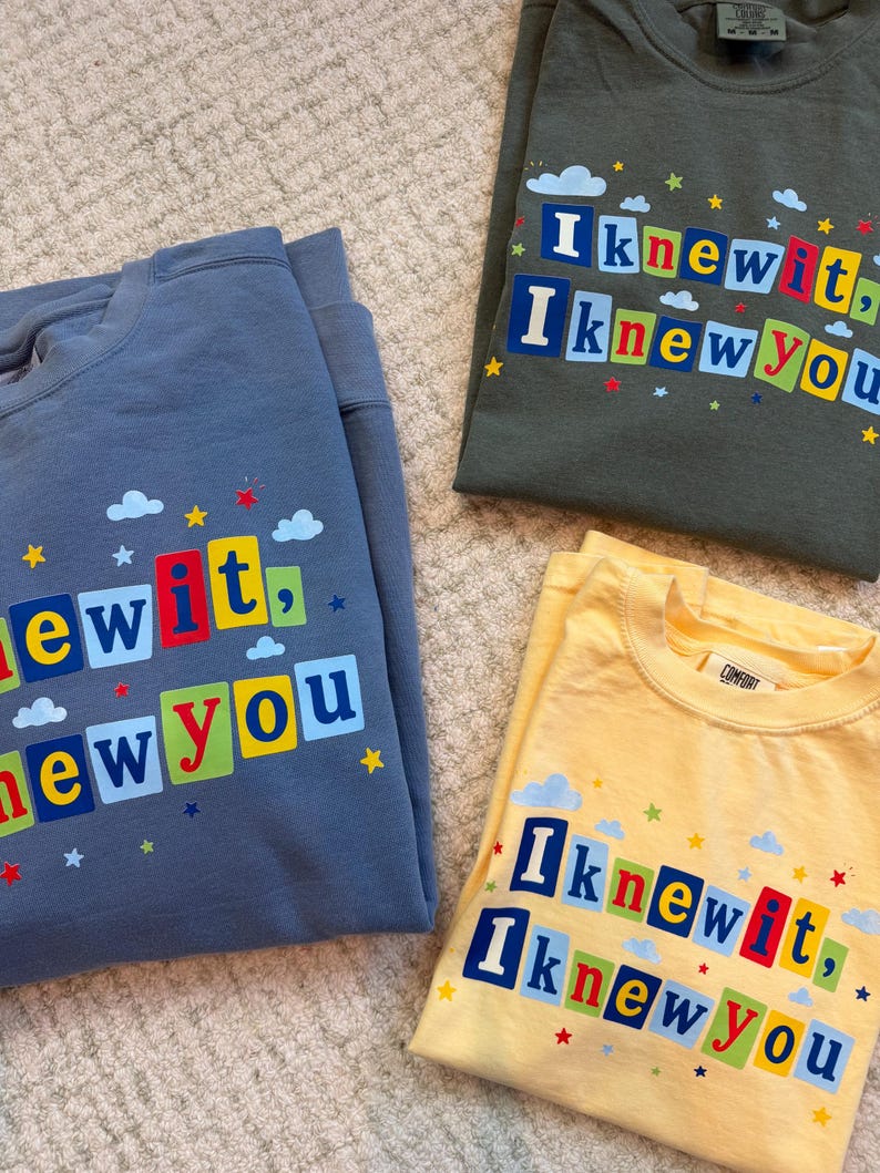 Op de afbeelding: Drie shirts met ronde hals in blauw, olijfgroen en geel. Elk shirt heeft een grafische print met de tekst "I knew it, I knew you" in kleurrijke blokletters, omringd door kleine blauwe wolken en rode, gele en blauwe sterren.