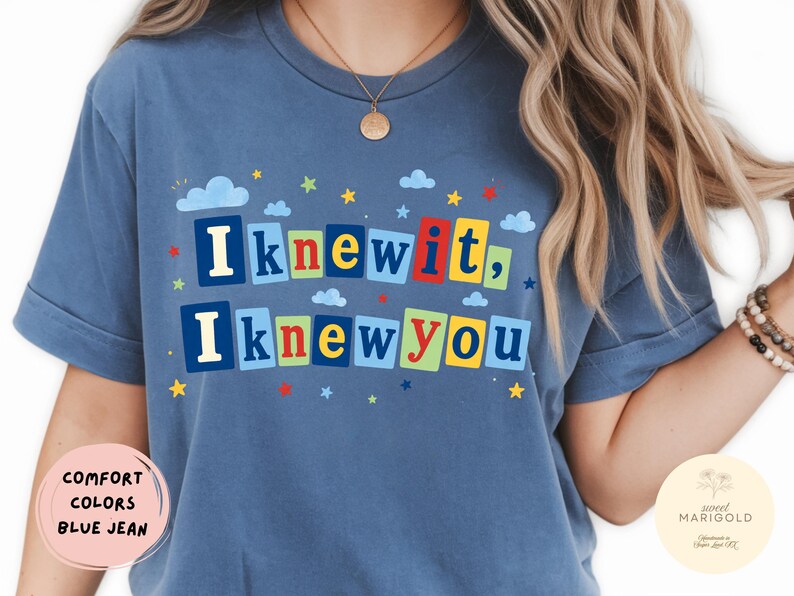 Op de afbeelding: Een blauw T-shirt met korte mouwen en de tekst "I knew it, I knew you" in kleurrijke blokletters. Het ontwerp is versierd met kleine blauwe wolkjes en veelkleurige sterretjes die rond de tekst verspreid zijn. Het shirt heeft een ronde hals.