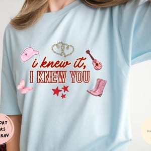 May include: A light blue t-shirt featuring the red text "i knew it, I KNEW YOU" surrounded by graphics of a pink cowboy hat, two intertwined ropes, a pink acoustic guitar, a pink butterfly, three red stars, and a pair of pink cowboy boots.