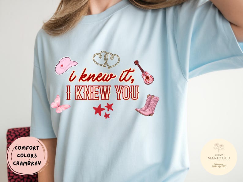 May include: A light blue t-shirt featuring the red text "i knew it, I KNEW YOU" surrounded by graphics of a pink cowboy hat, two intertwined ropes, a pink acoustic guitar, a pink butterfly, three red stars, and a pair of pink cowboy boots.