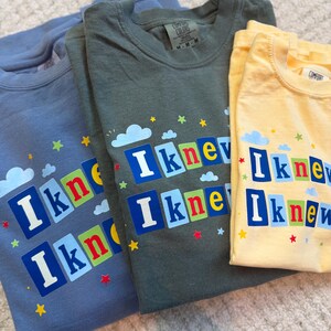 Op de afbeelding: Drie t-shirts in blauw, olijfgroen en geel, elk voorzien van de tekst "I knew" in witte letters op kleurrijke vierkante blokken. Het ontwerp bevat kleine blauwe wolken en verspreide rode, gele en groene sterren rondom de tekst.