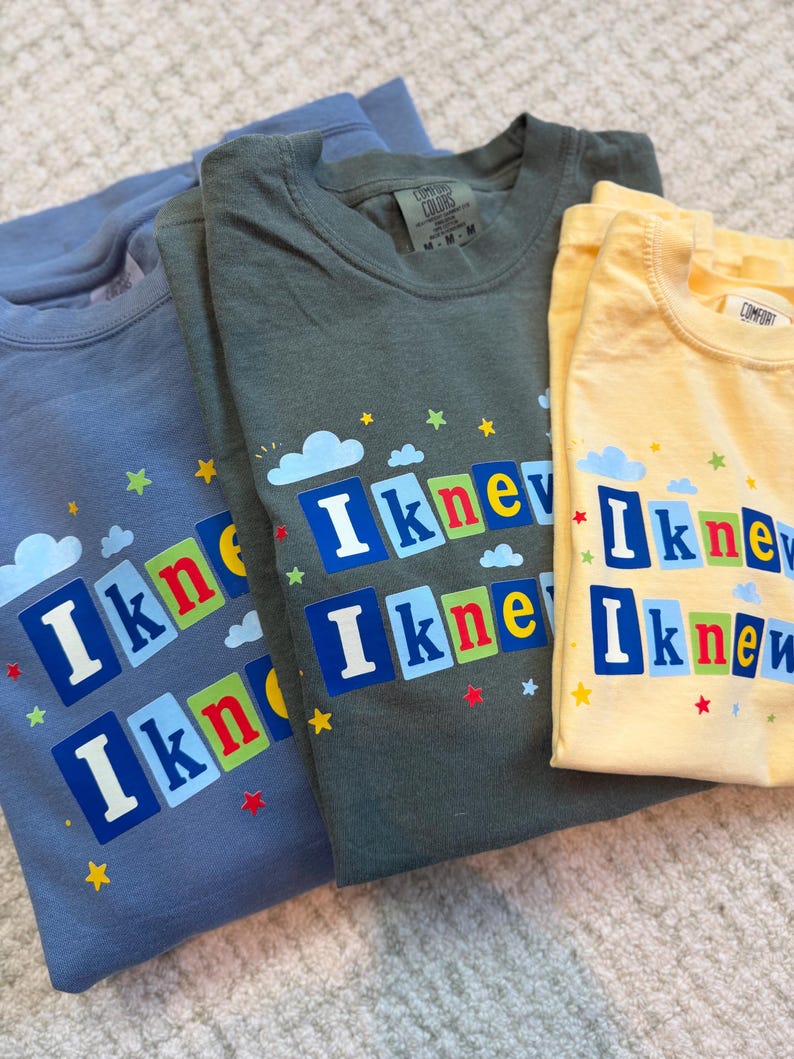 Op de afbeelding: Drie t-shirts in blauw, olijfgroen en geel, elk voorzien van de tekst "I knew" in witte letters op kleurrijke vierkante blokken. Het ontwerp bevat kleine blauwe wolken en verspreide rode, gele en groene sterren rondom de tekst.