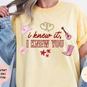 May include: A butter yellow t-shirt featuring the red text "i knew it, I KNEW YOU" surrounded by illustrations of a pink cowboy hat, two intertwined rope hearts, a pink acoustic guitar, a pink butterfly, three red stars, and pink cowboy boots.