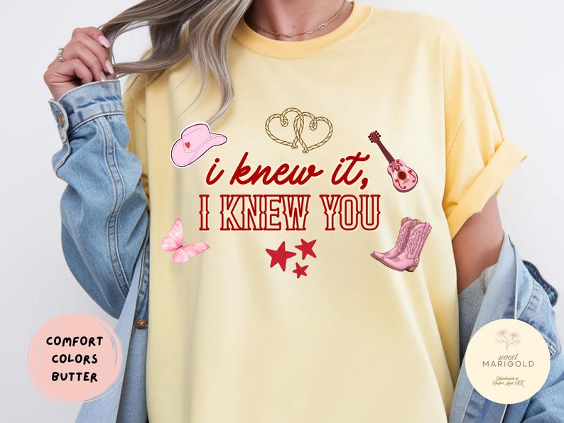 May include: A butter yellow t-shirt featuring the red text "i knew it, I KNEW YOU" surrounded by illustrations of a pink cowboy hat, two intertwined rope hearts, a pink acoustic guitar, a pink butterfly, three red stars, and pink cowboy boots.