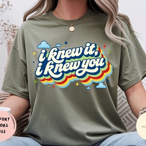 May include: A moss green short-sleeve t-shirt featuring the text "i knew it, i knew you" in a dark blue script font. The text is layered over a multi-colored wavy graphic in red, yellow, and blue, accented by small blue clouds and stars.