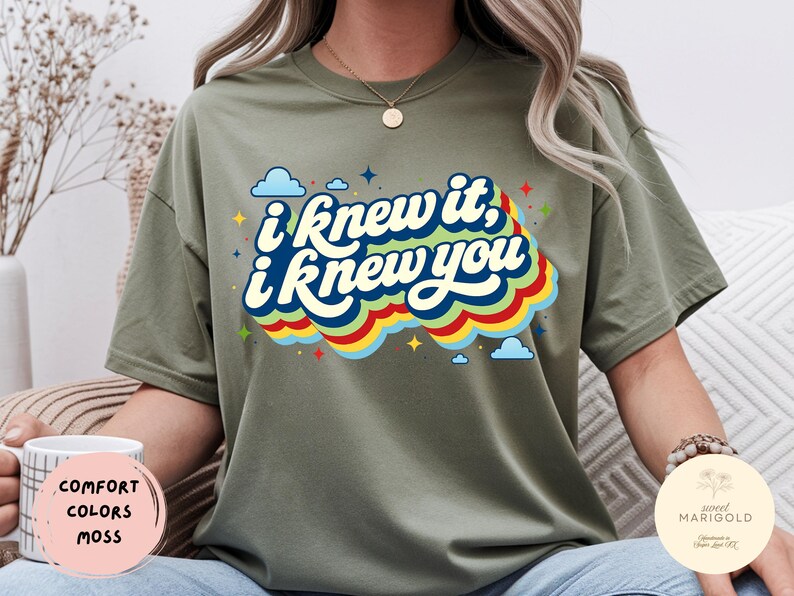 May include: A moss green short-sleeve t-shirt featuring the text "i knew it, i knew you" in a dark blue script font. The text is layered over a multi-colored wavy graphic in red, yellow, and blue, accented by small blue clouds and stars.