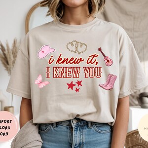 May include: An ivory t-shirt featuring the red text "i knew it, I KNEW YOU" surrounded by illustrations of a pink cowboy hat, a rope heart, a pink guitar, a pink butterfly, three red stars, and a pair of pink cowboy boots on the front.
