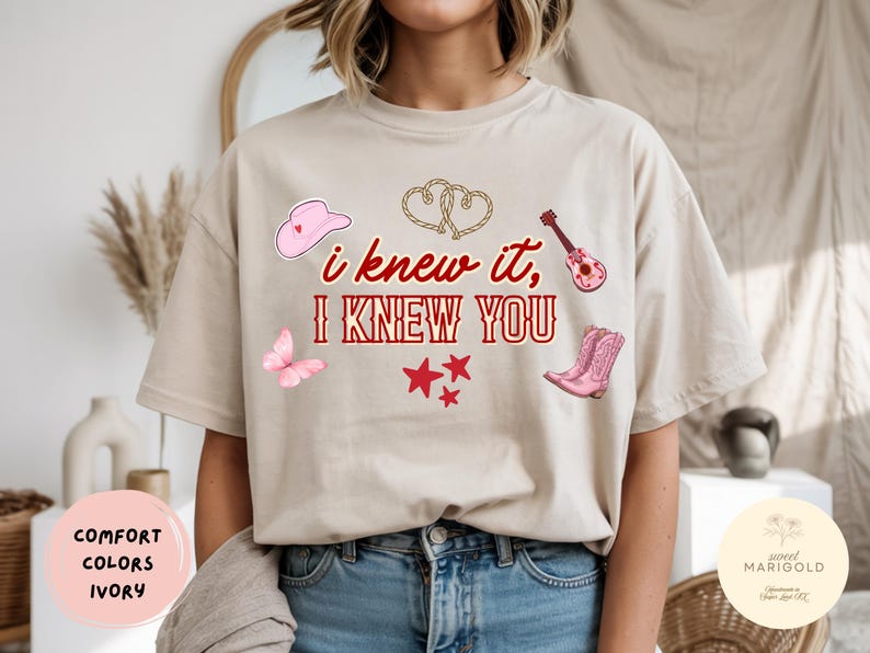 May include: An ivory t-shirt featuring the red text "i knew it, I KNEW YOU" surrounded by illustrations of a pink cowboy hat, a rope heart, a pink guitar, a pink butterfly, three red stars, and a pair of pink cowboy boots on the front.