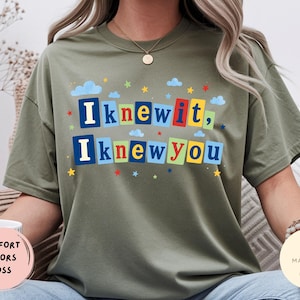 Op de afbeelding: Een olijfgroen t-shirt met korte mouwen en de tekst "Iknewit, Iknewyou" in een kleurrijke blokletterstijl. De letters staan op rechthoekige achtergronden in blauw, rood, geel en groen, geaccentueerd door kleine blauwe wolkjes en sterretjes.