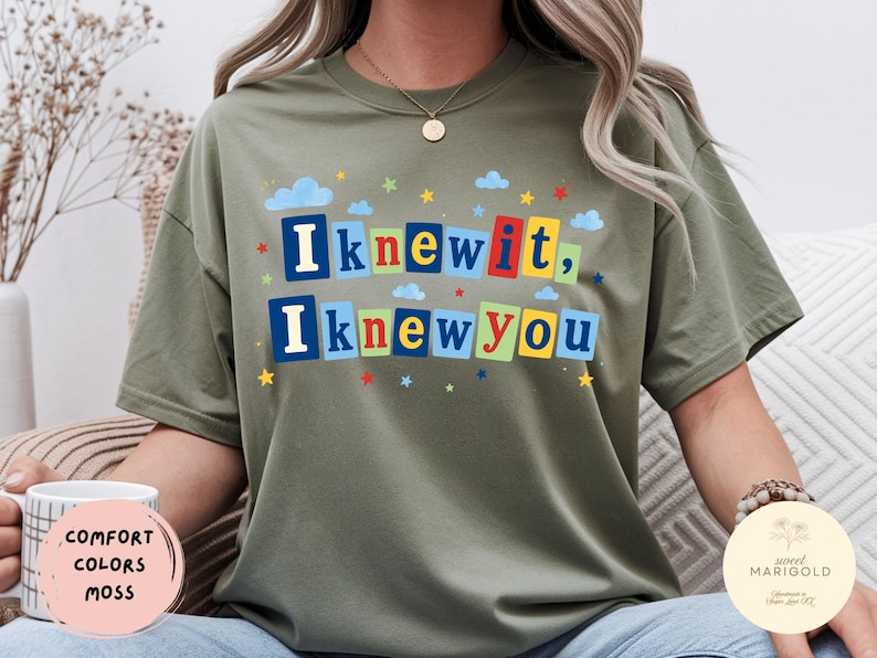 Op de afbeelding: Een olijfgroen t-shirt met korte mouwen en de tekst "Iknewit, Iknewyou" in een kleurrijke blokletterstijl. De letters staan op rechthoekige achtergronden in blauw, rood, geel en groen, geaccentueerd door kleine blauwe wolkjes en sterretjes.
