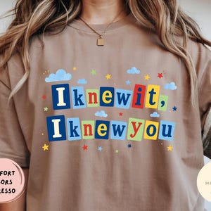 Op de afbeelding: Een bruin t-shirt met de tekst "Iknewit, Iknewyou" in kleurrijke blokletters. Het ontwerp is versierd met kleine blauwe wolken en diverse veelkleurige sterren die rond de tekst zijn verspreid, wat zorgt voor een speelse en levendige grafische uitstraling.